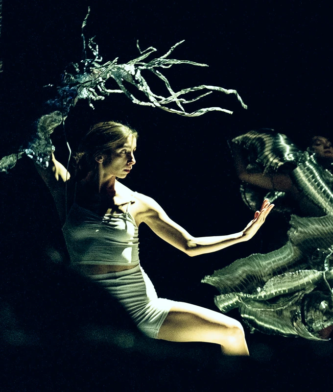 Tancerka w białym stroju pozuje dynamicznie na ciemnej scenie z metalicznymi rekwizytami